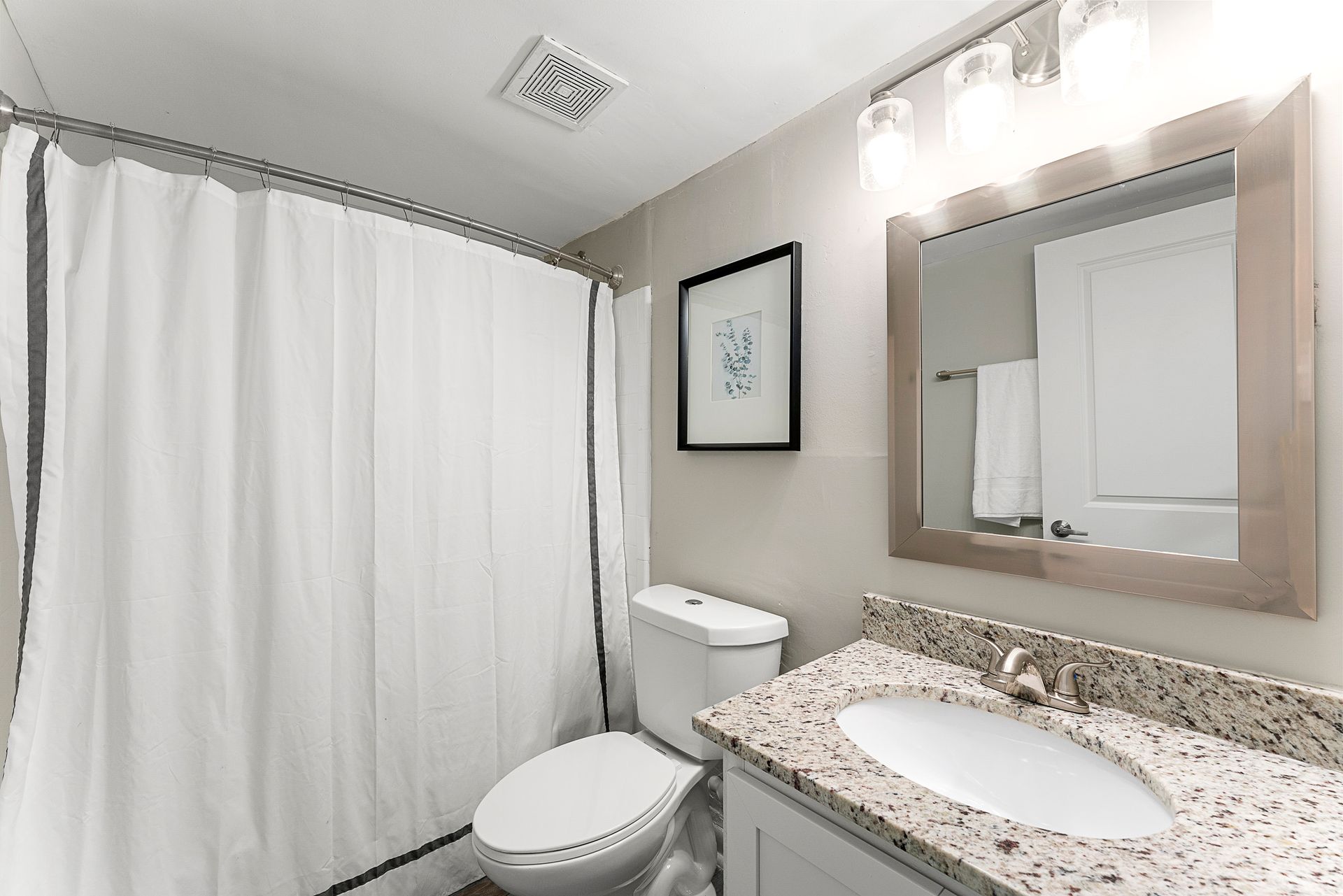 Bright bathroom with shower curtain, toilet, granite vanity, and large mirror.
