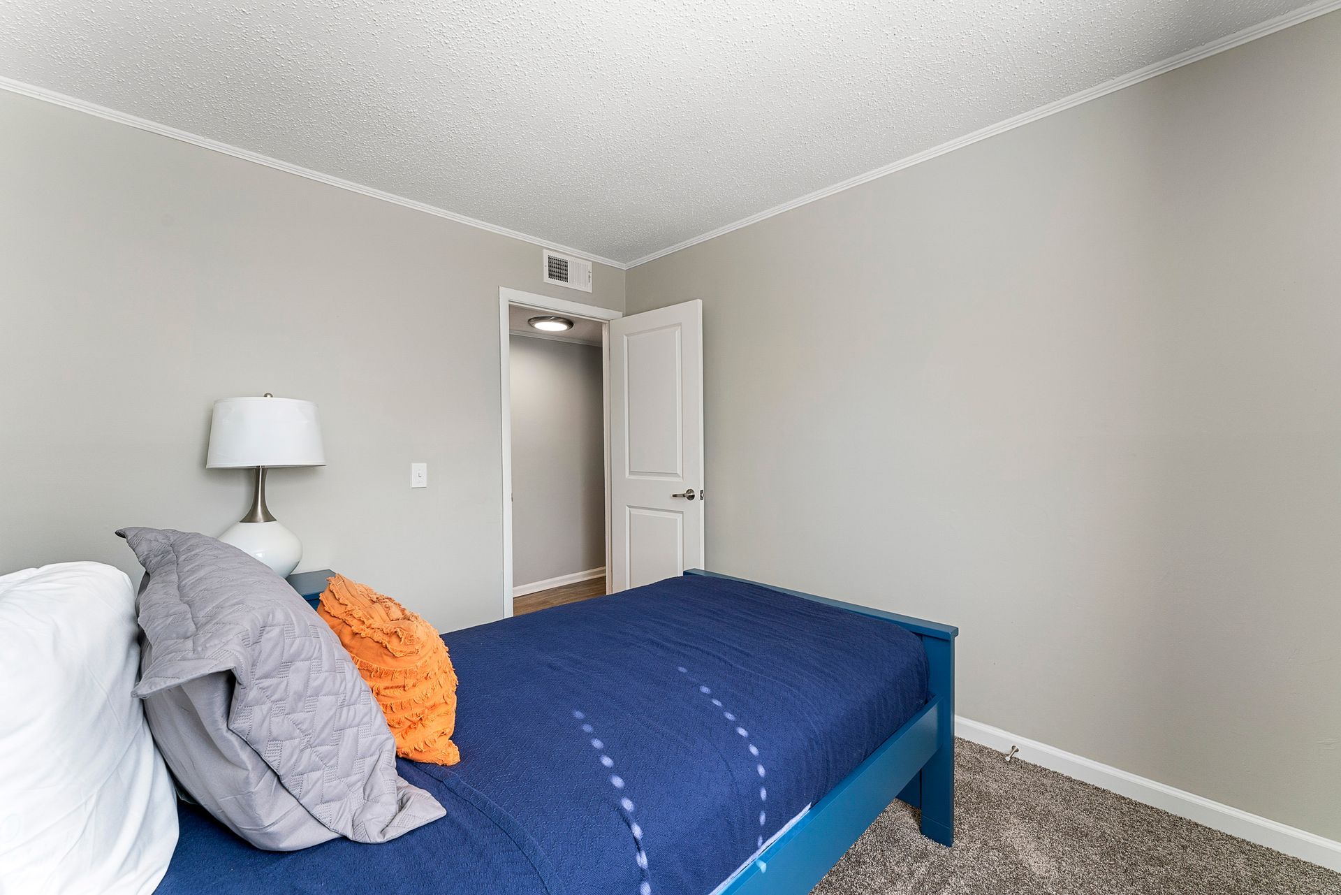 Small bedroom with a blue bedspread, gray pillows, beige walls, and an open door.