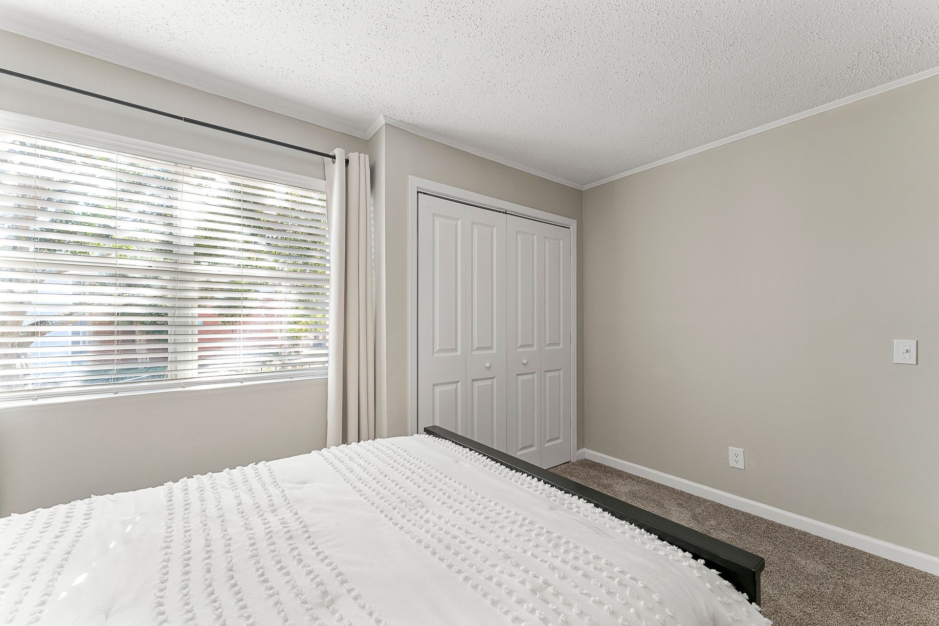 Bright bedroom with white bedspread, window blinds, and closed closet doors.