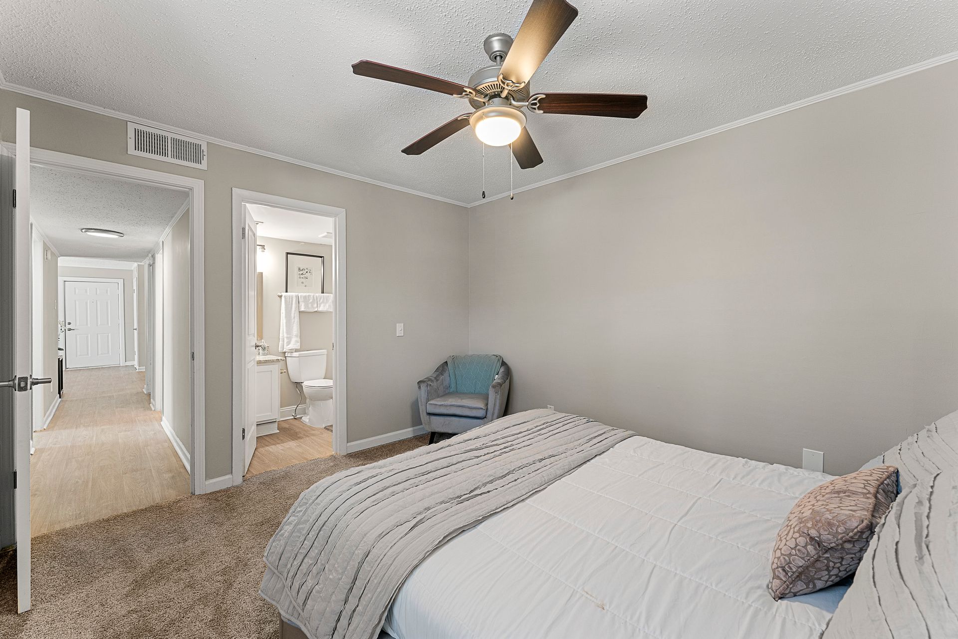 Bright bedroom with gray walls, ceiling fan, bed, chair, and open doorway to a bathroom and hall.