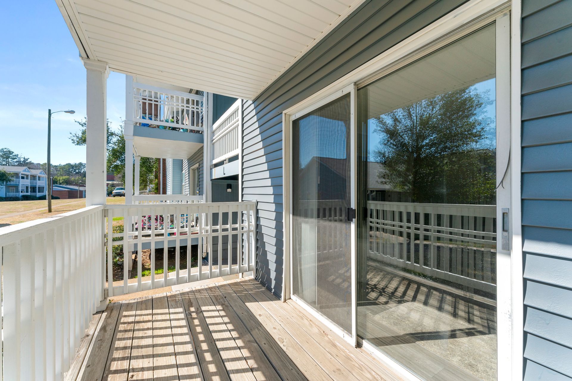 Covered apartment balcony with white railing, blue siding, and sliding glass doors in bright sunlight