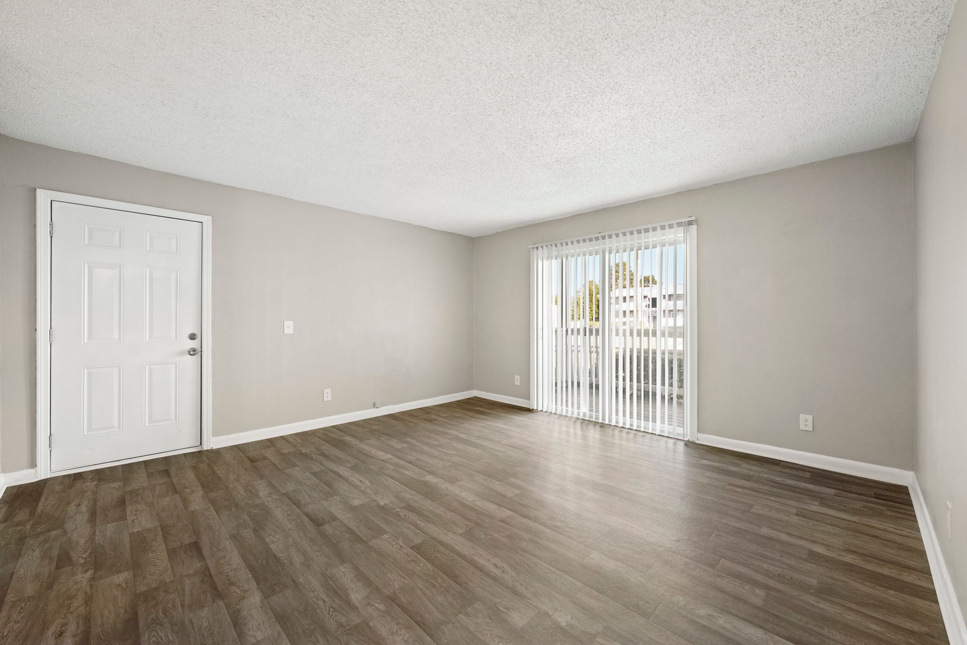 Empty beige living room with wood flooring, white door, and sliding glass door with vertical blinds