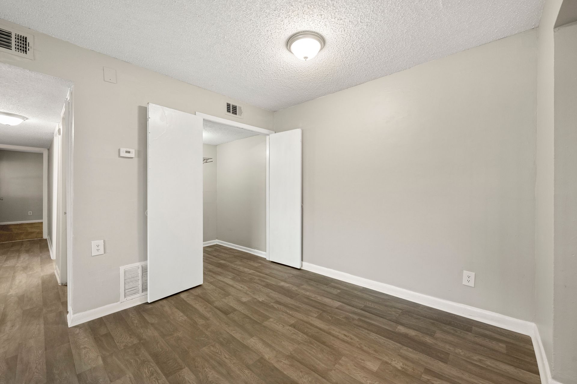 Empty white room with wood flooring, open closet doors, and a hallway visible to the left