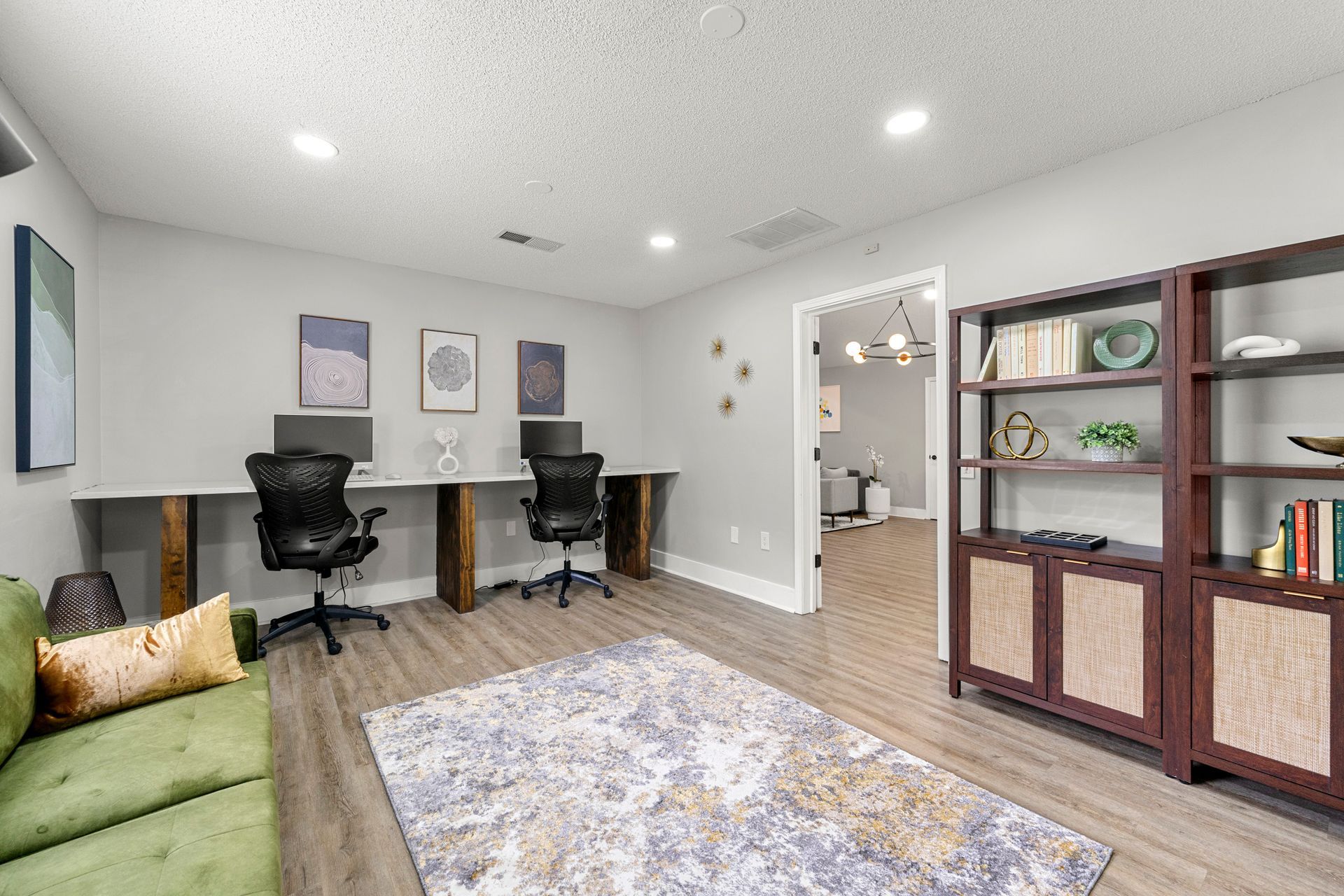Modern home office with two desks, black chairs, a green sofa, and a bookshelf on wood floors.