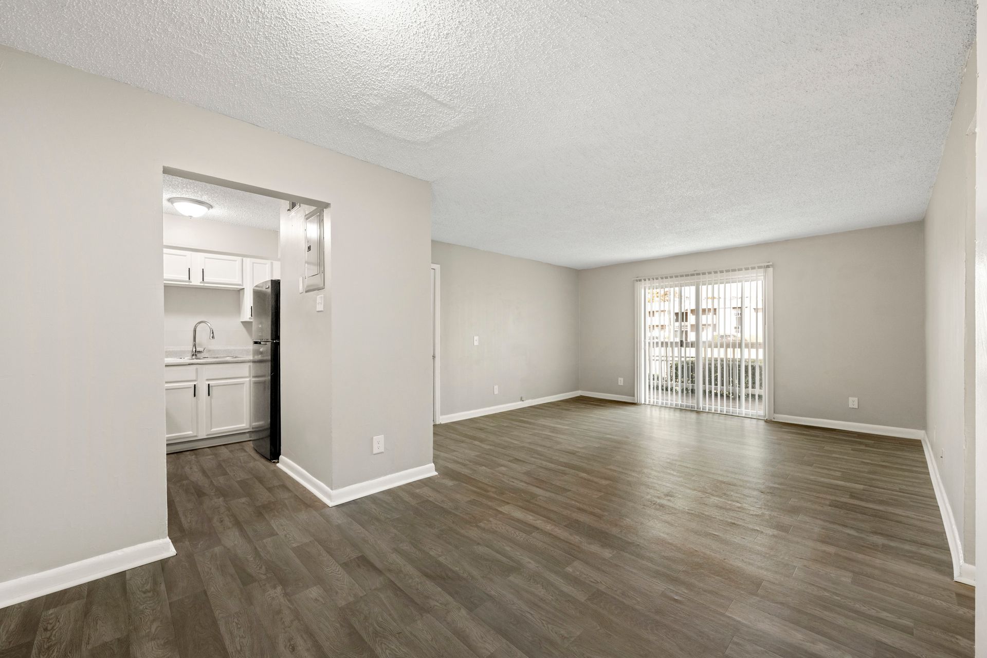 Empty beige living room with wood flooring, open kitchen entry, and sliding glass doors to a balcony.