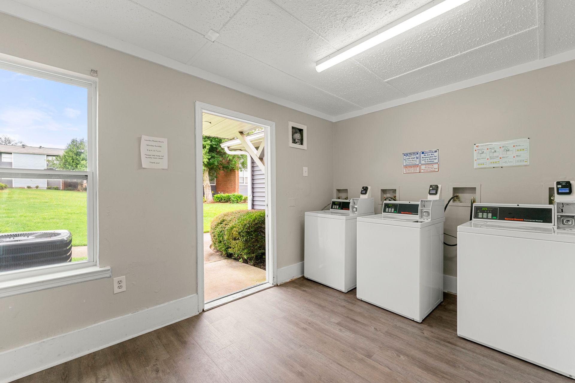 Bright laundry room with white appliances, gray walls, wood floor, and open door to a sunny yard