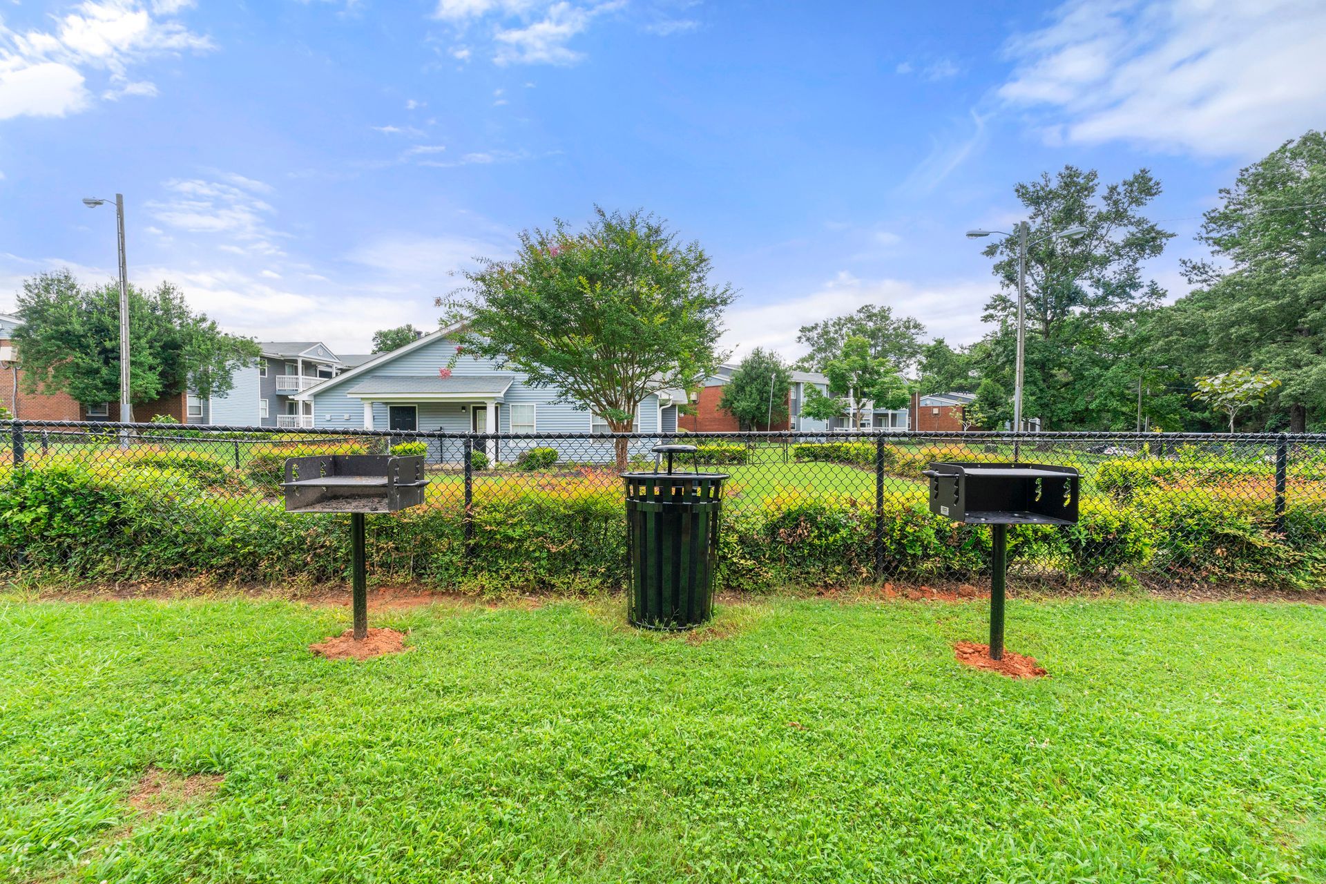 Grassy park with two black grills, a fence, trees, and houses under a bright blue sky.