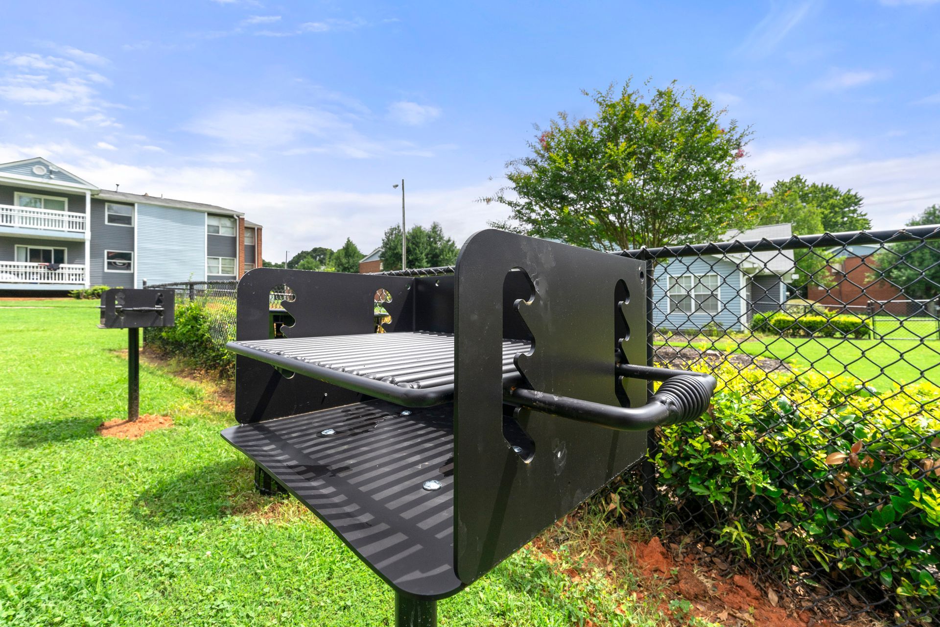 Outdoor black barbecue grill beside a chain-link fence on a grassy lawn near apartment buildings