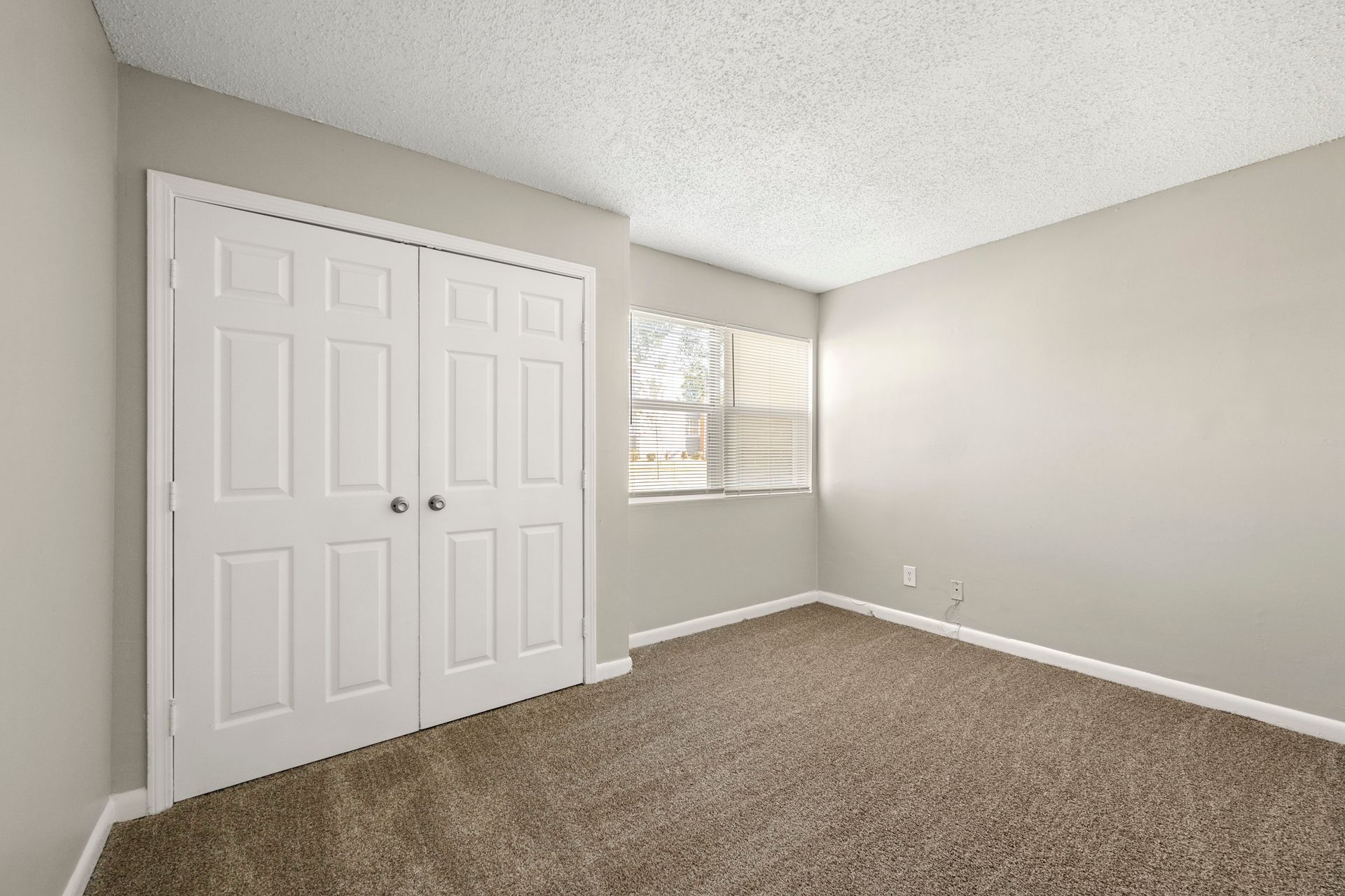 Empty beige bedroom with carpet, white closet doors, and a window with blinds