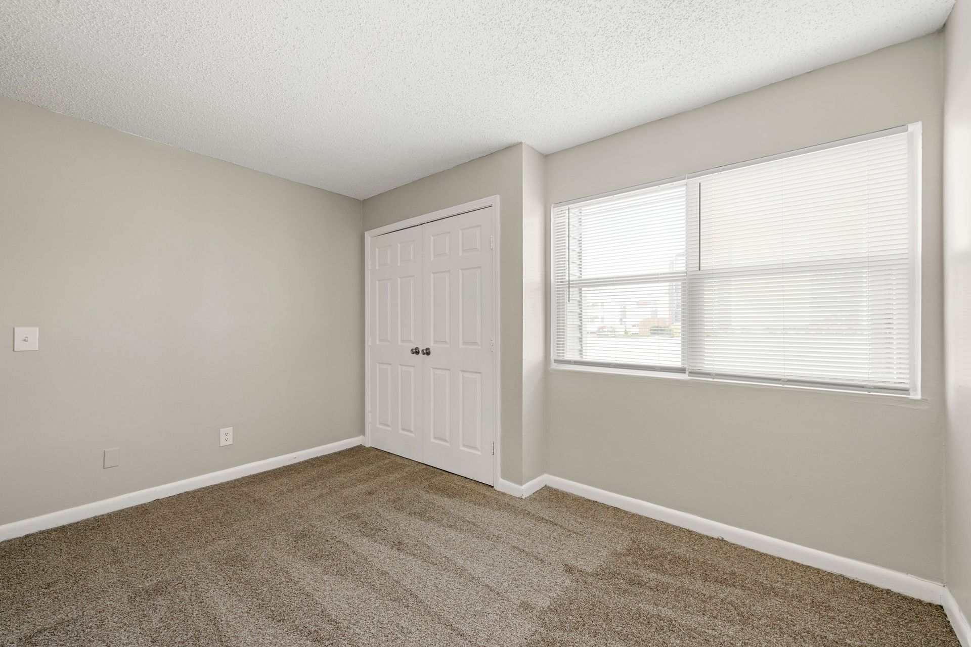 Empty beige bedroom with carpet, white closet doors, and a window with blinds.