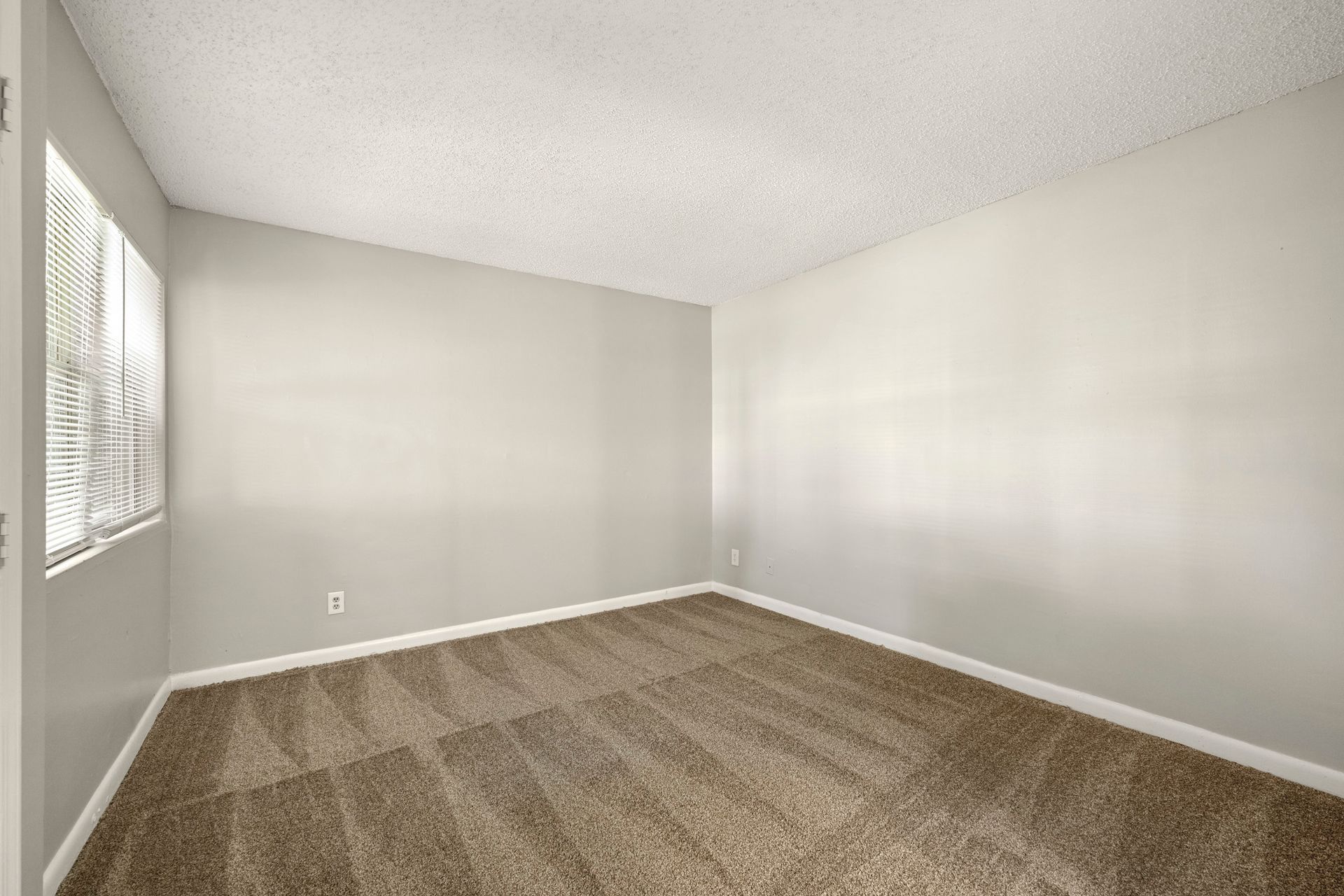 Empty beige carpeted room with white walls and a window on the left.