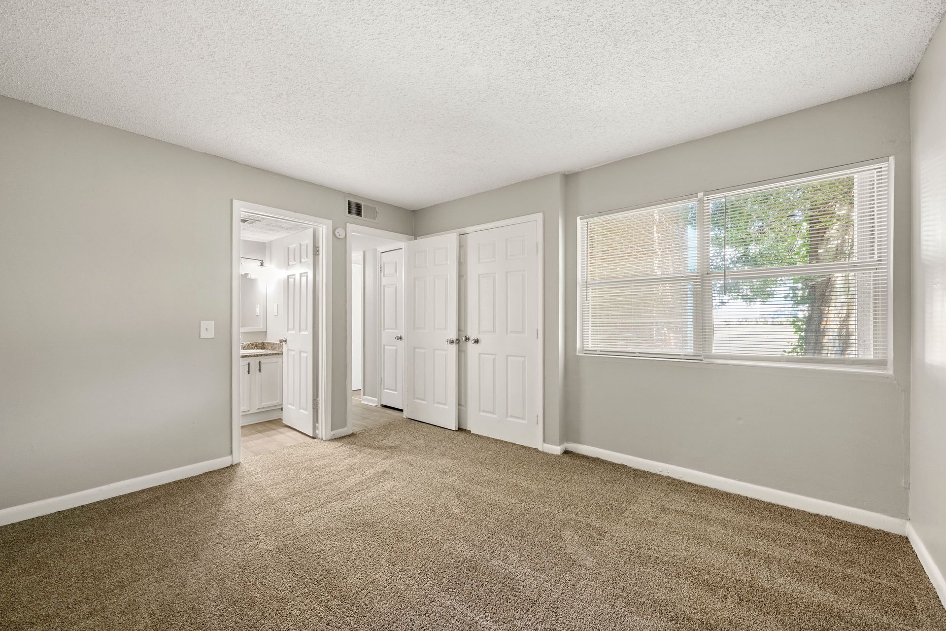 Empty beige bedroom with carpet, white doors, and a window with blinds