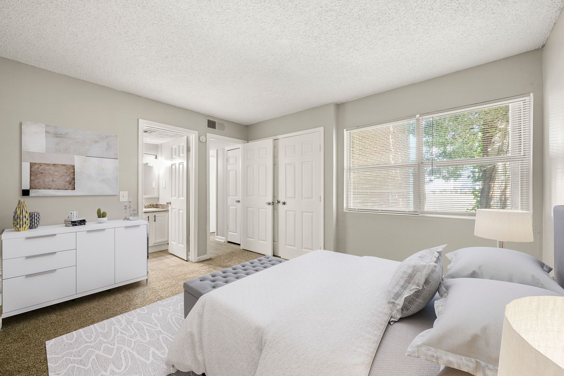 Bright bedroom with a bed, white dresser, closet doors, and a large window with blinds
