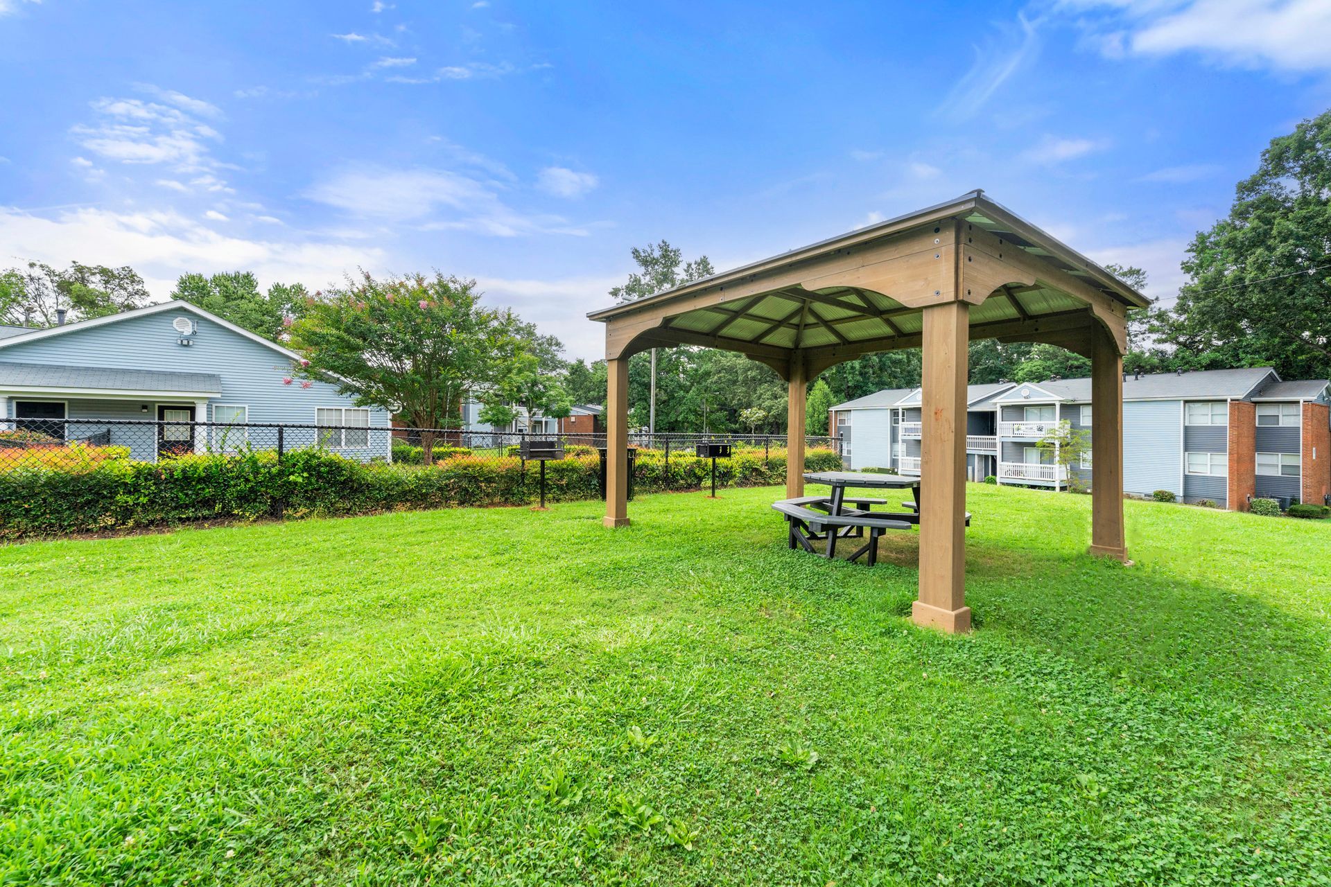 Grassy park with a wooden gazebo and picnic table under a blue sky, bordered by houses and hedges