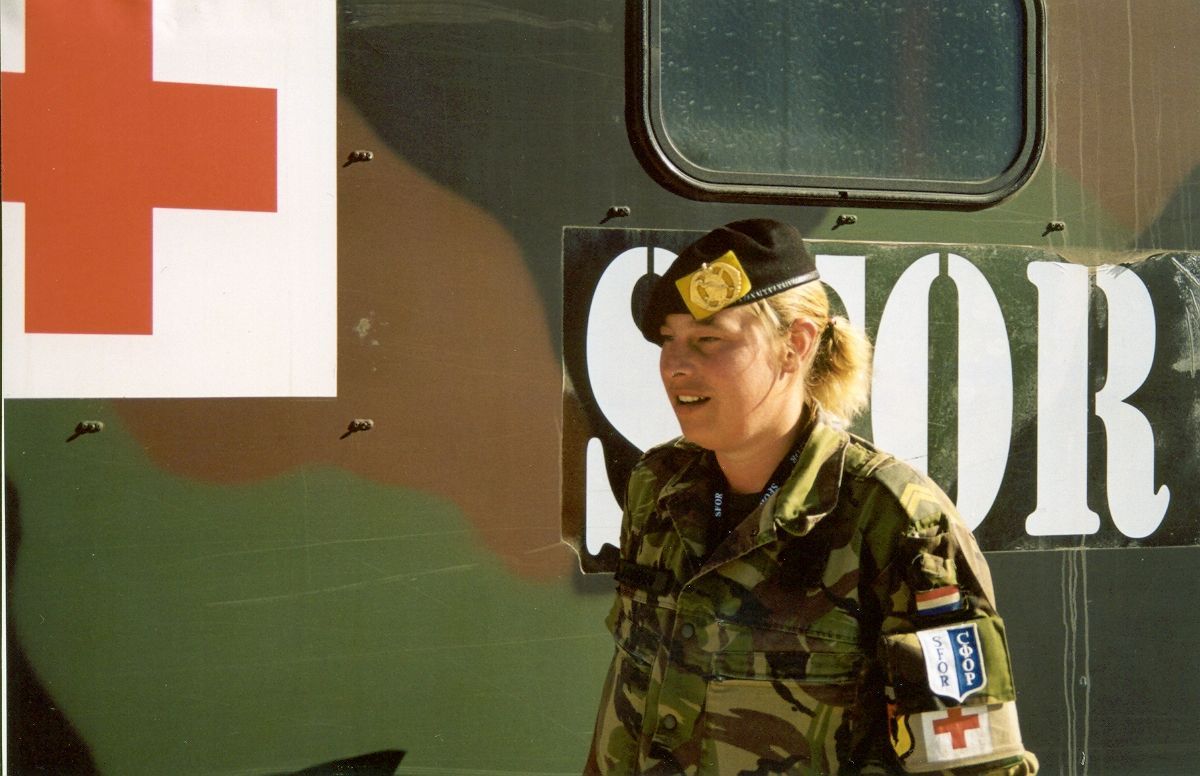 Een persoon in militair uniform met een rood kruis-embleem staat voor een voertuig met het opschrift