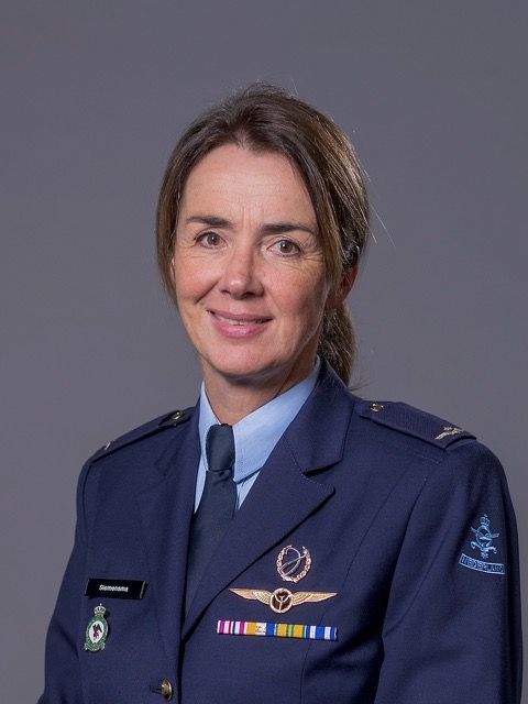 Een militair officier in een blauw uniform met medailles en insignes poseert voor een formeel portret tegen een grijze achtergrond.