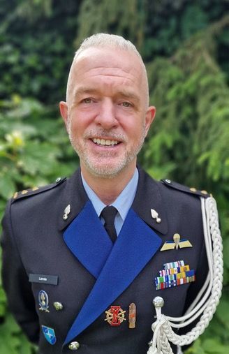 Een glimlachende militaire officier in een formeel donkerblauw uniform met medailles en helmtekens, staand voor groen gebladerte.
