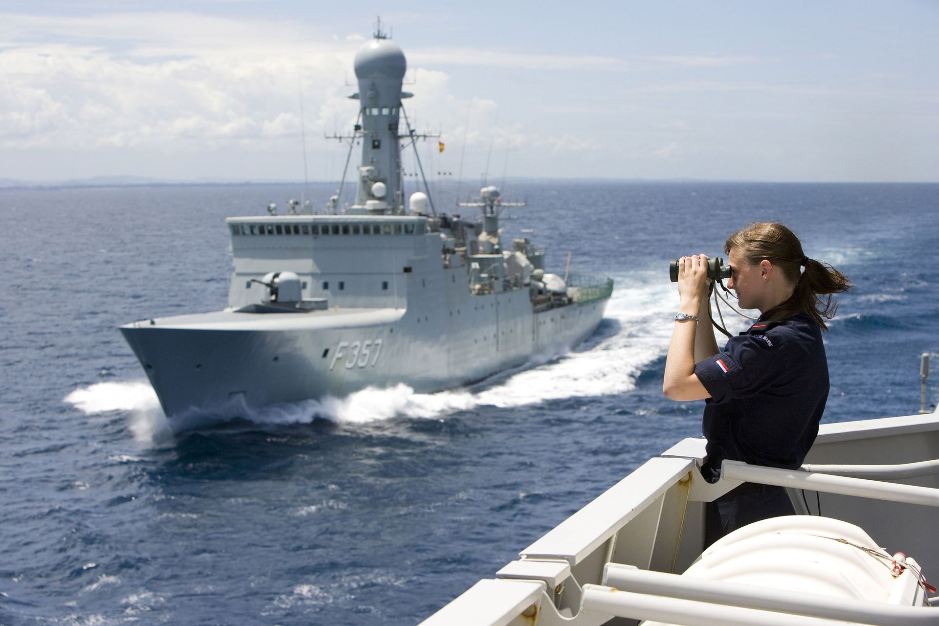 Iemand gebruikt een verrekijker om een ​​grijs marineschip te bekijken dat op open zee vaart.