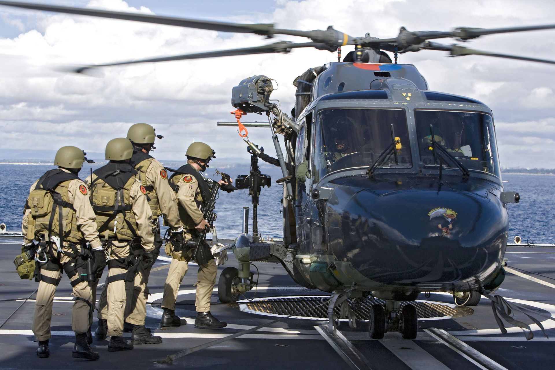 Vier soldaten in beige gevechtskleding staan ​​op het dek van een schip, vlakbij een donkere militaire helikopter waarvan de rotorbladen draaien.