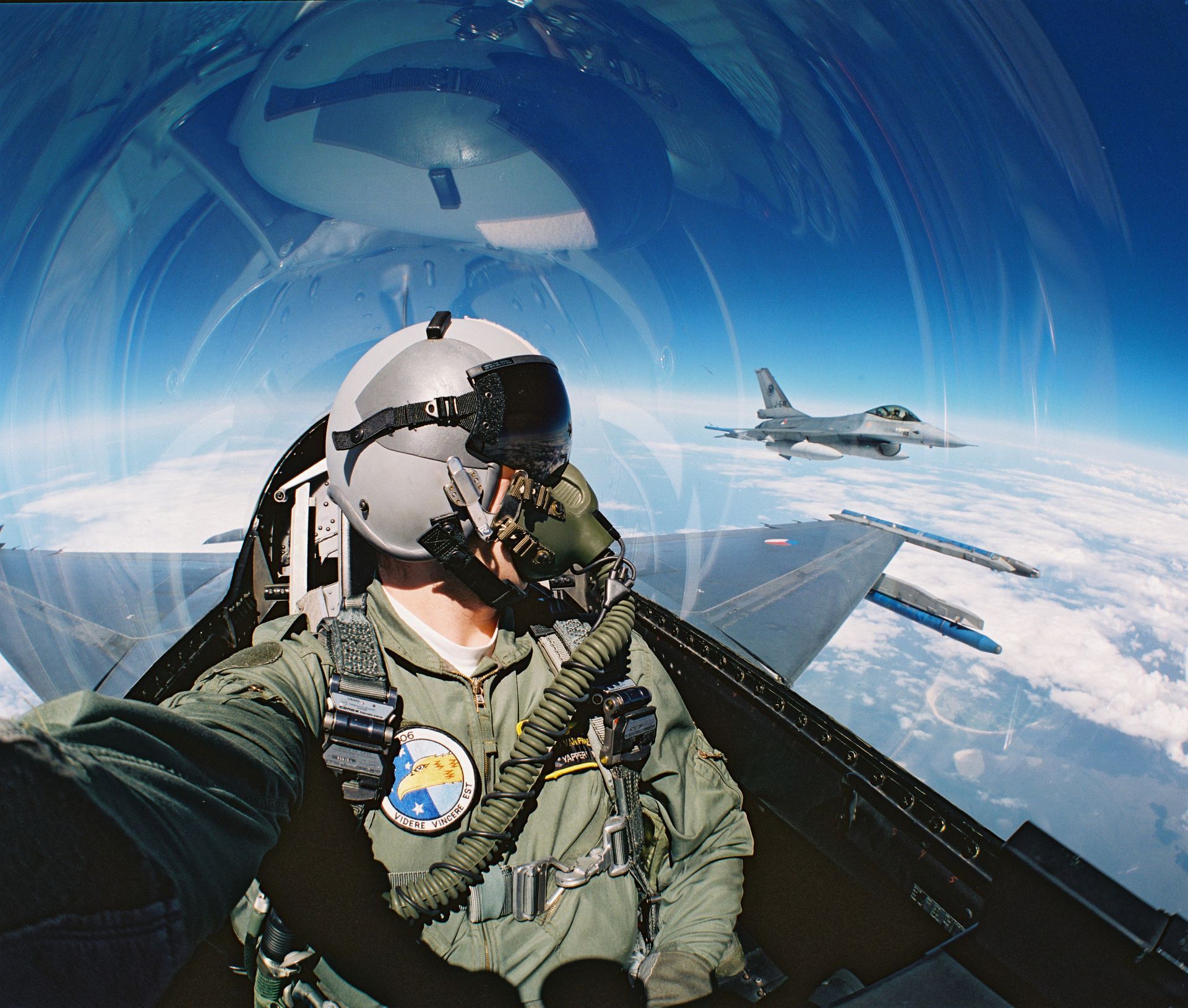 Een gevechtspiloot maakt een selfie vanuit de cockpit terwijl hij hoog boven de wolken naast een andere F-16 vliegt.