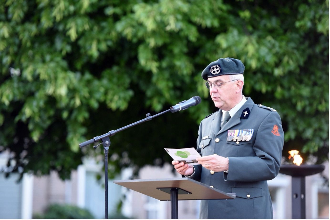 Een militair in uniform staat buiten achter een podium en leest voor uit een document in een microfoon.