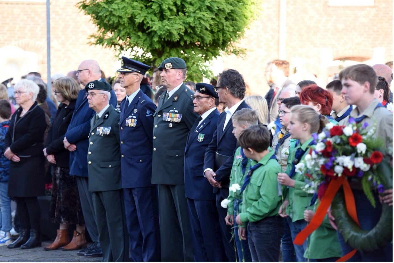 Militairen en verkenners staan in een plechtige formatie in de open lucht tijdens een herdenkingsceremonie.
