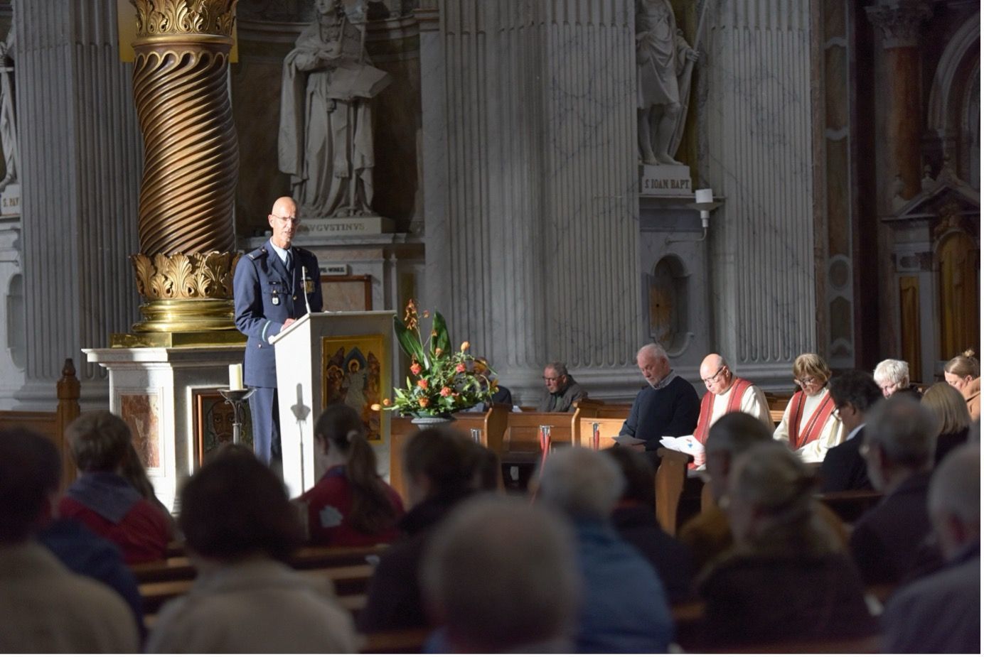 Een persoon in een formeel militair uniform staat op een podium in een kerk en spreekt het publiek toe
