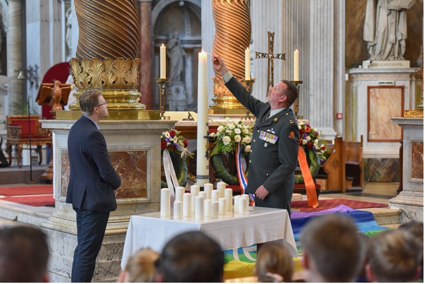 Een persoon in militair uniform steekt een hoge kaars aan in een kerk, terwijl een andere persoon toekijkt