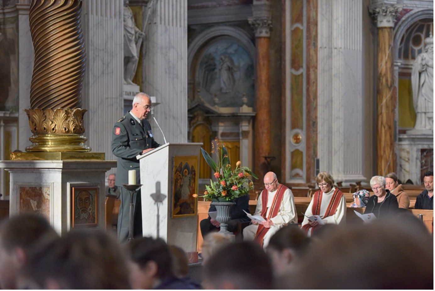 Een functionaris in uniform spreekt vanaf een lessenaar in een rijkversierde kerk, terwijl de aanwezigen in de buurt zitten.