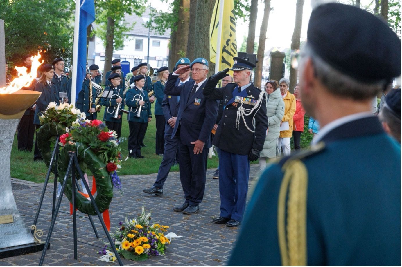 Veteranen in uniform brengen een saluut tijdens een herdenkingsceremonie bij een brandende eeuwige vlam en bloemenkransen in een park.