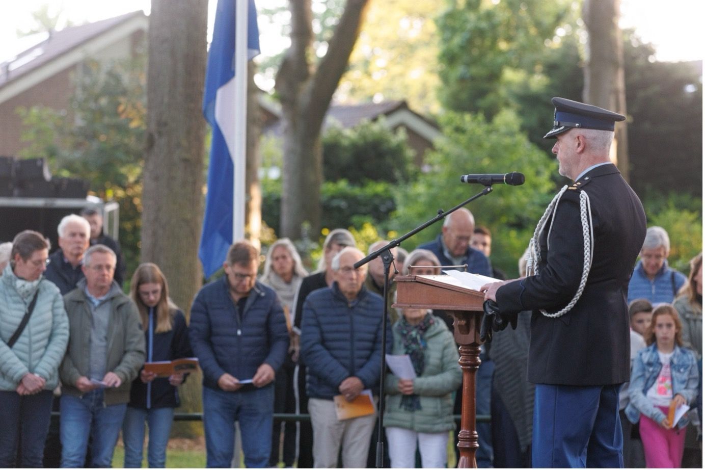 Een agent in uniform spreekt vanaf een podium tijdens een herdenkingsdienst in de openlucht, met een menigte op de achtergrond.