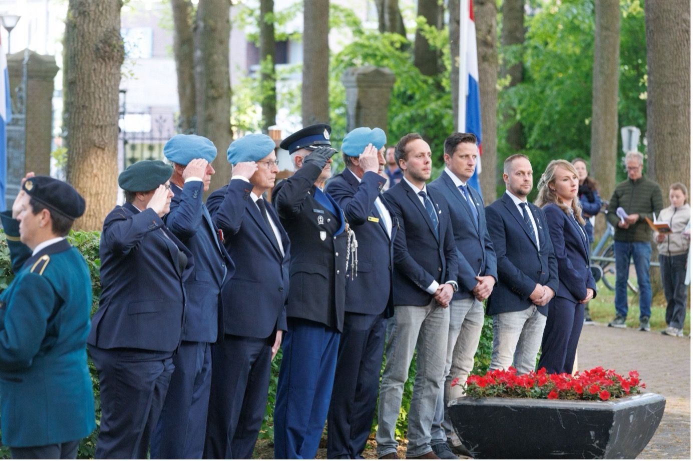 Een rij mensen in militaire uniformen en galakleding brengt een saluut tijdens een herdenkingsceremonie in de openlucht.