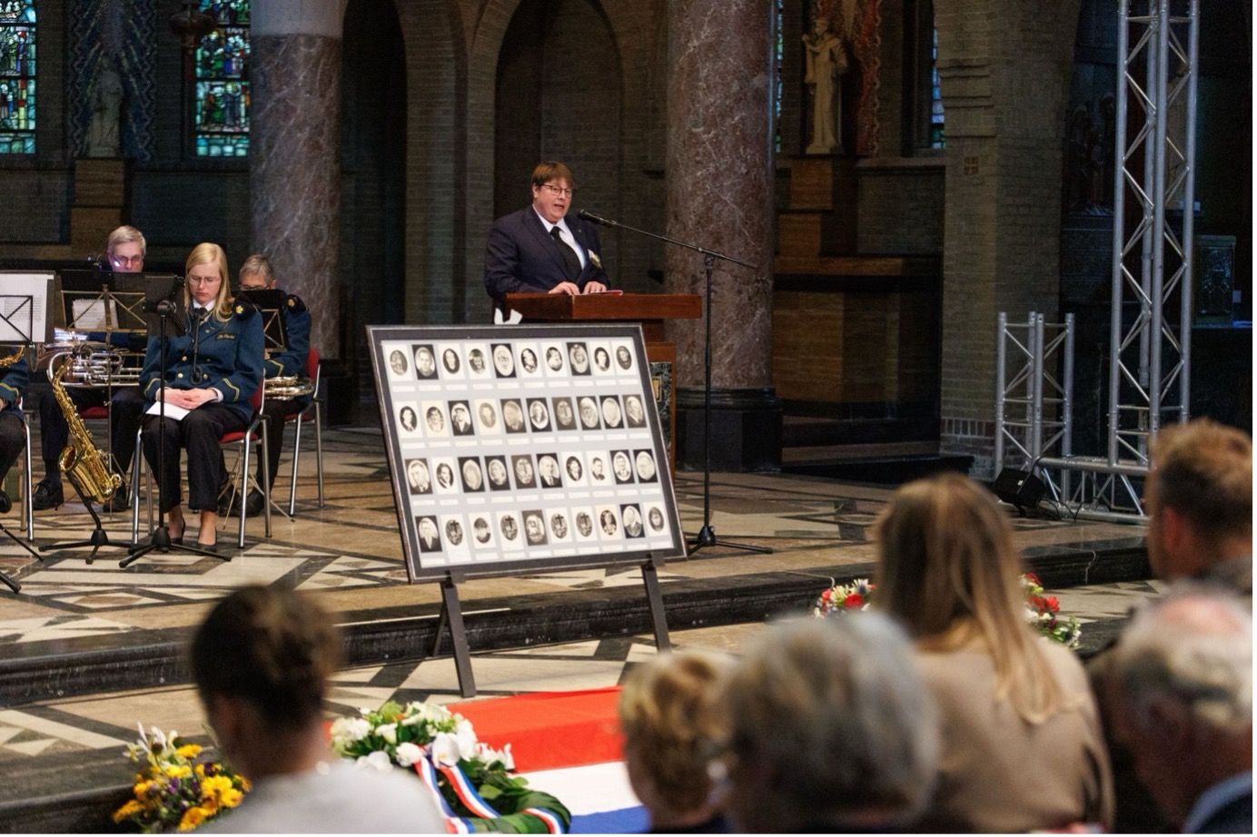 Een spreker staat op een podium in een kerk, tegenover een gedenkplaat met portretten van aanwezigen op de voorgrond.