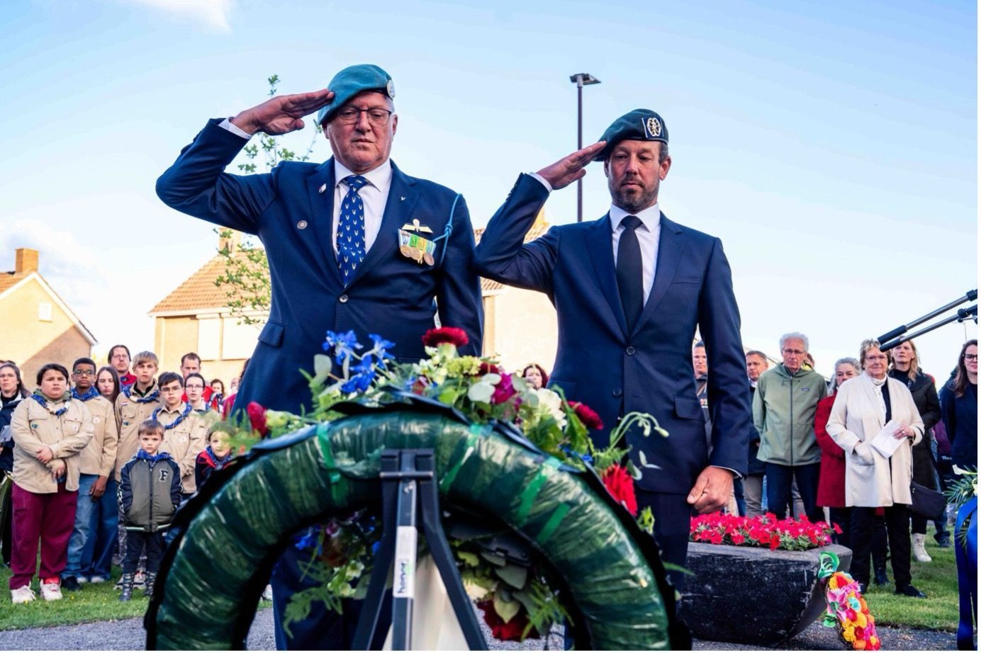 Twee personen in galakleding en groene baretten brengen een saluut aan een bloemenkrans
