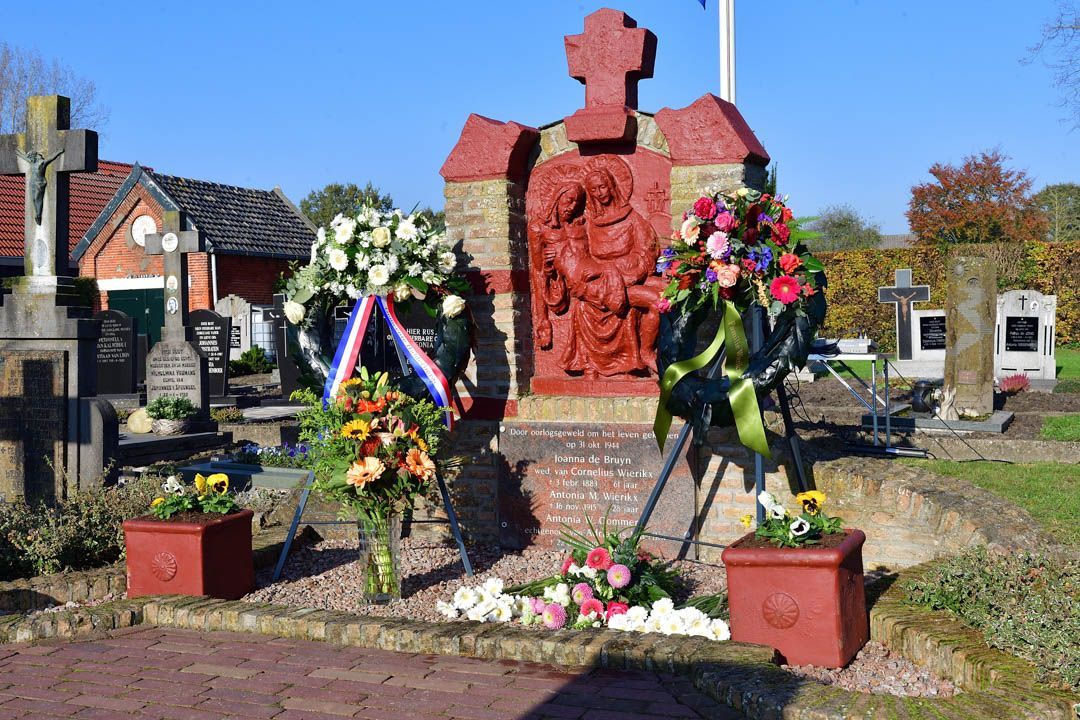 Een gedenkteken met een rood religieus reliëf en kruis, versierd met bloemenkransen en plantenbakken, in een begraafplaatsomgeving.