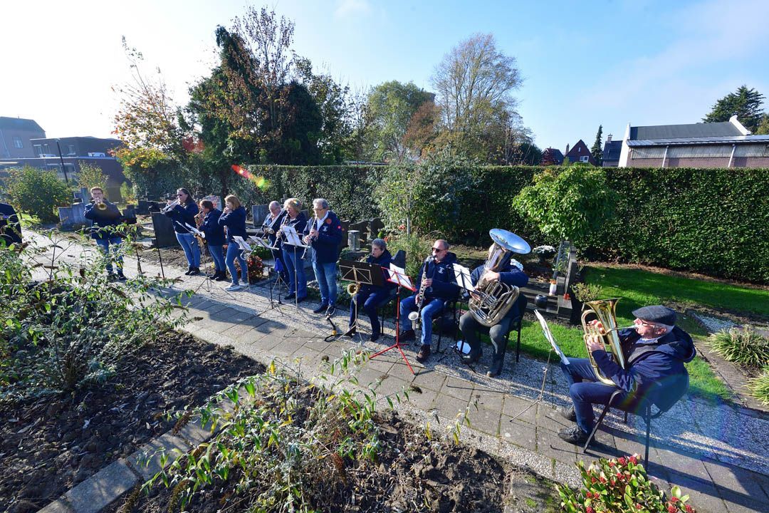 Een fanfare speelt instrumenten zittend en staand langs een pad in een park of op een begraafplaats op een zonnige dag.