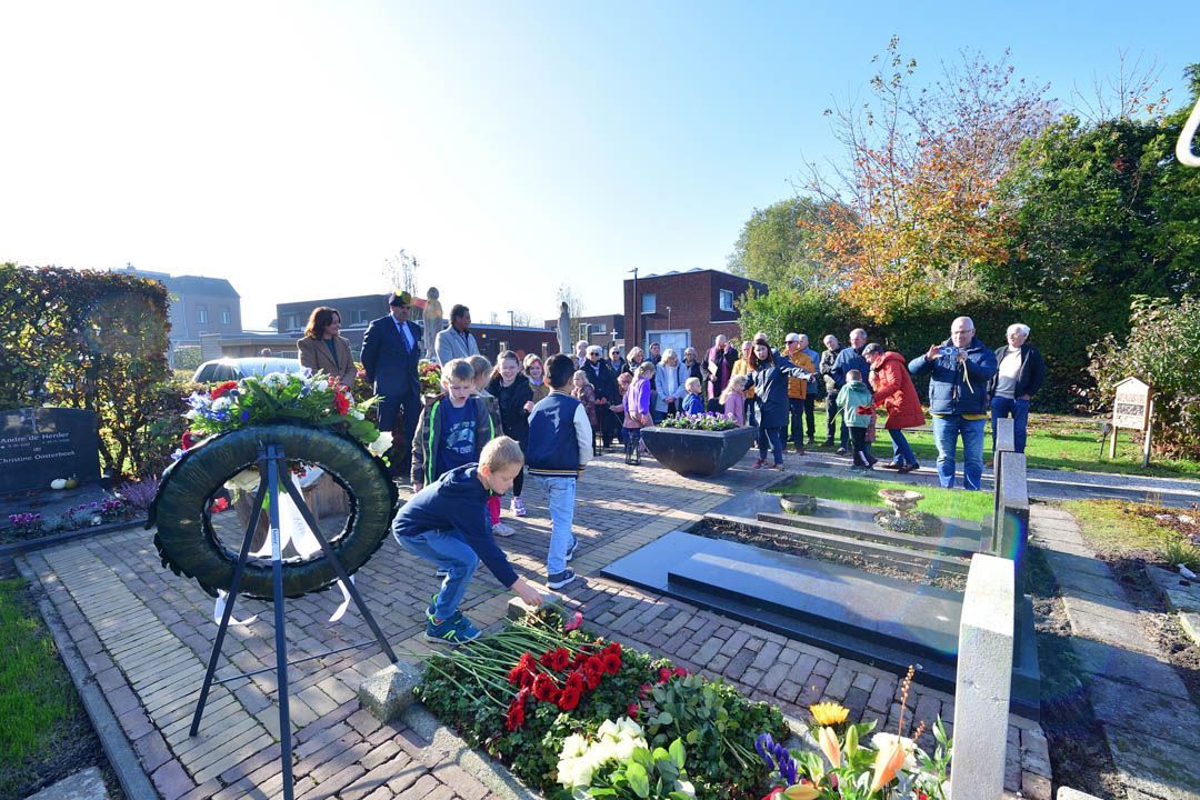 Mensen verzamelden zich op een begraafplaats om kransen en bloemen op een graf te leggen tijdens een zonnige herdenkingsdienst.