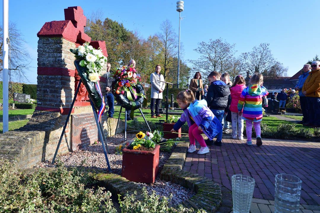Mensen verzamelen zich bij een stenen oorlogsmonument in de openlucht, waar een kind bloemen in een bloempot aan de voet van het monument plaatst.