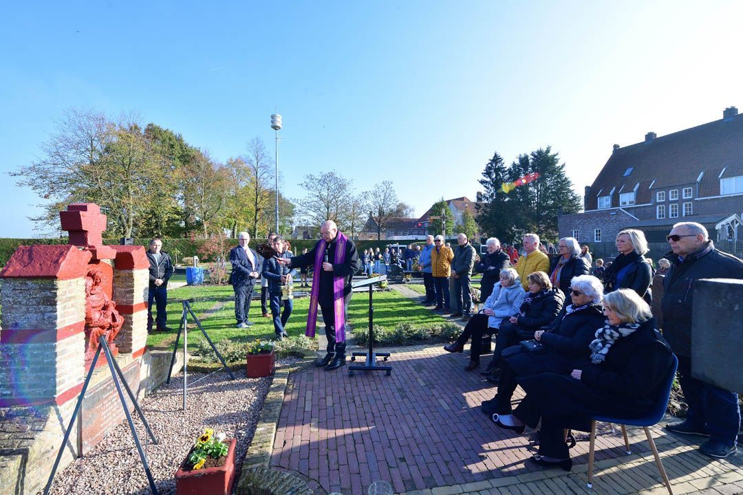 Een groep mensen verzamelt zich buiten voor een stenen monument tijdens een herdenkingsdienst die wordt geleid door iemand in een geestelijke gewaad.