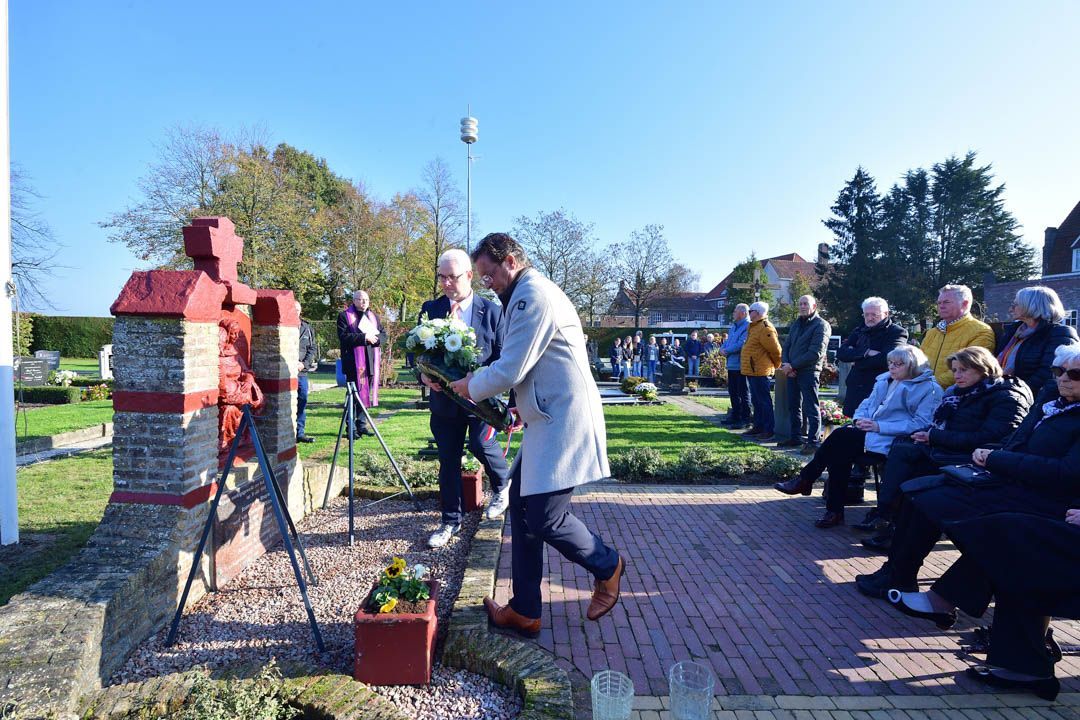 Een persoon in een lichte jas legt een bloemenkrans bij een stenen monument, terwijl een kleine groep mensen buiten toekijkt.
