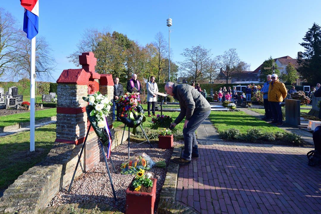 Een man legt bloemen neer bij een stenen monument op een begraafplaats op een zonnige dag, met een vlag die er vlakbij wappert.