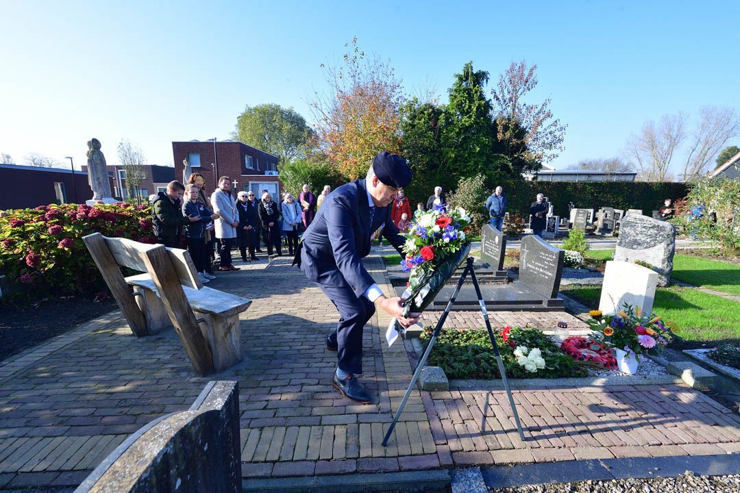 Een persoon in een donker pak legt een bloemenkrans op een graf tijdens een herdenkingsceremonie in de openlucht, met toeschouwers in de buurt.