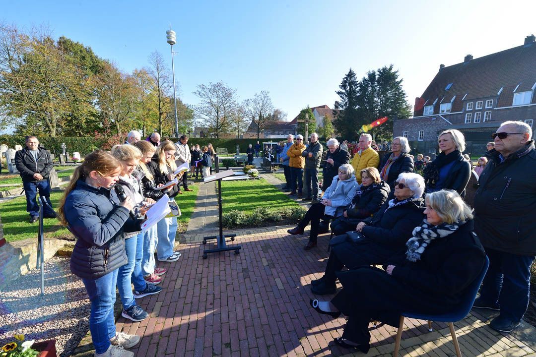 Een groep mensen staat buiten op een begraafplaats voor een herdenkingsdienst, waarbij sommigen voorlezen uit papieren.