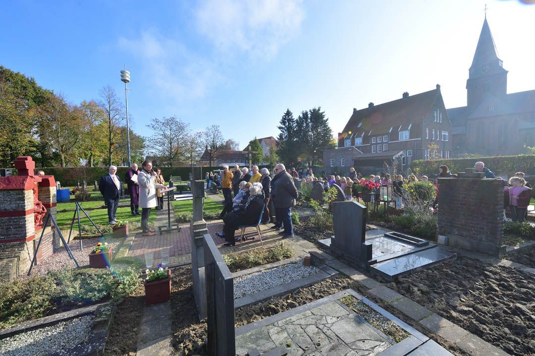 Een groep mensen staat op een begraafplaats vlakbij een kerk en luistert naar een spreker tijdens een herdenkingsdienst in de openlucht.