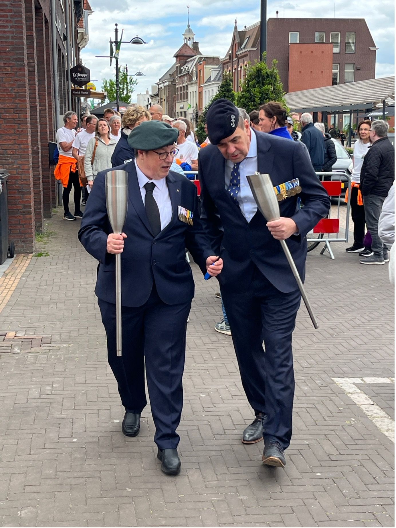 Twee personen in donkere pakken en baretten lopen naast elkaar over een straat, elk met een ceremoniële fakkel in de hand.