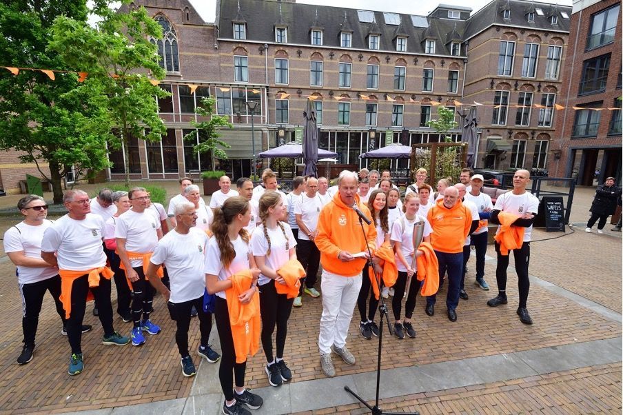 Een groep mensen in witte overhemden en oranje jassen op een plein terwijl een man in een microfoon spreekt.