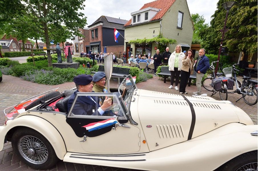 Twee mensen in een crèmekleurige oldtimer met een fakkel op een stadsplein, omringd door toeschouwers.