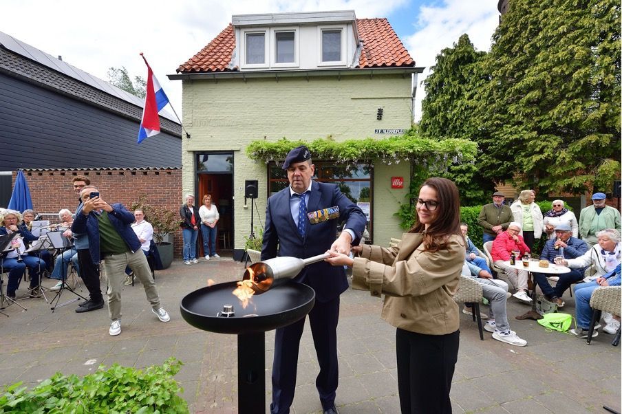 Een persoon in een pak en een persoon in een jasje steken een ceremoniële vlam aan met een fakkel op een binnenplaats.