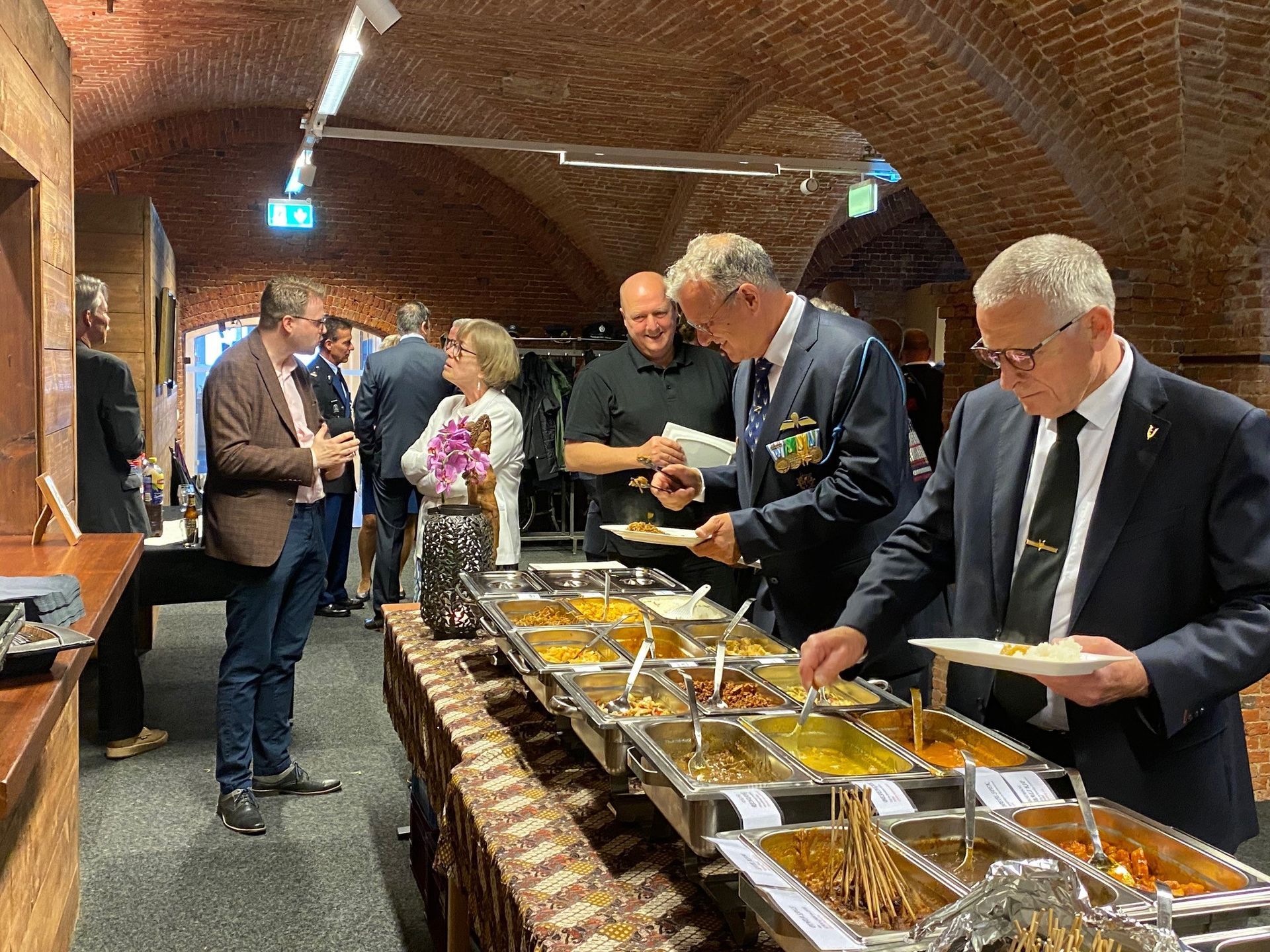Een groep mensen in zakelijke kleding verzamelt zich bij een buffet in een eetzaal met zichtbare bakstenen bogen.