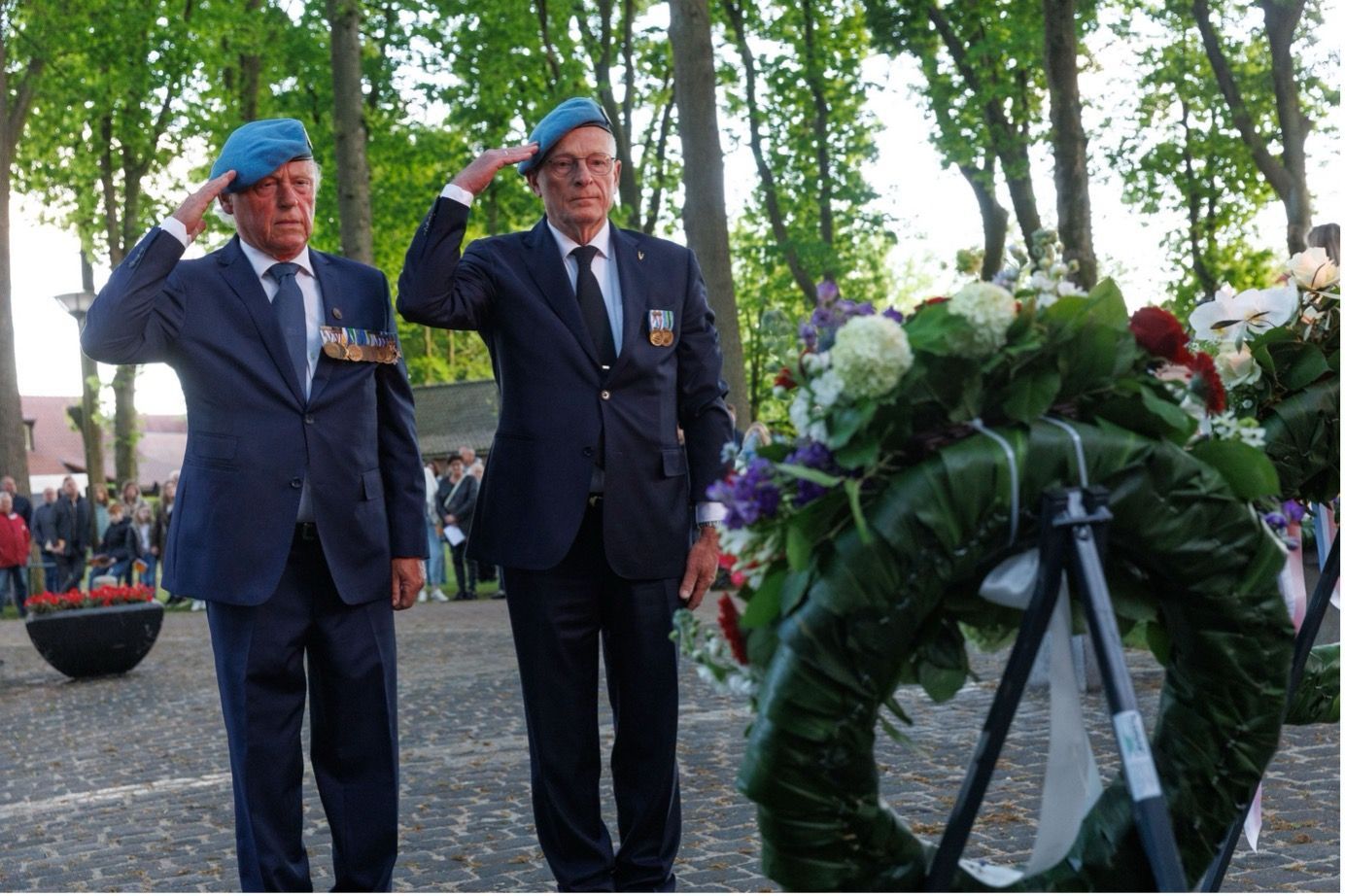 Twee mannen in pak en met blauwe baretten staan in een park en brengen een saluut aan een herdenkingskrans